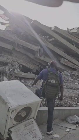 A man in a blue shirt, jeans, and a backpack walks towards a collapsed building. Several people in hard hats and vests are visible amongst the rubble, apparently engaged in rescue or recovery efforts. A washing machine lies on its side in the foreground. The sky is bright and hazy, suggesting it is daytime.