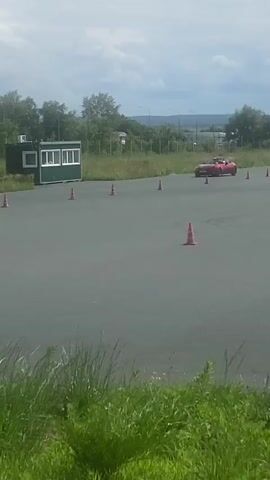 A red convertible sports car is shown driving around a paved track marked with orange cones. The car is seen from various angles as it navigates the course. In the background, there is a small green building with white windows, trees, and a cloudy sky. Tall grass is visible in the foreground. The car appears to be performing some sort of driving exercise, possibly a slalom or autocross event. The driver is visible inside the car, wearing a helmet.