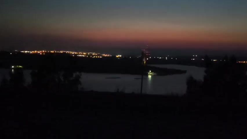 This is a static, low-light, medium-shot video clip showing a dark landscape at twilight. In the foreground, silhouetted trees obscure the view of the ground. In the middle ground, a body of water, possibly a river, curves through the scene. In the background, a city skyline is visible with numerous lights twinkling against a twilight sky that transitions from dark blue at the top to shades of pink and orange near the horizon. A tall, thin tower with red lights is visible near the center of the background, likely part of the city's infrastructure. The overall atmosphere is dark and somewhat hazy.