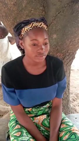 A young woman is sitting on a wooden bench under a tree. She has dark skin and is wearing a headscarf, a black and blue shirt, and a green skirt. She is looking down and has her hands folded in her lap. There is a man standing behind her and to the left, partially obscured by the tree trunk. The ground is covered in sand. The scene appears to be outdoors in a sandy area, possibly near a beach.