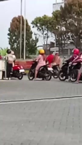 The video shows a gas station scene. Several people are present, some on motorcycles. Two individuals, one in a red uniform and another in a lighter colored outfit, are standing near a motorcycle. Another person in a pink outfit and a yellow helmet is riding a motorcycle. There are gas pumps visible, and a black car is parked. The environment appears to be outdoors, with trees and a building in the background. The lighting suggests it might be daytime.
