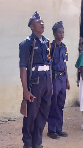 Two young men in matching navy blue uniforms, consisting of short-sleeved shirts, trousers, and garrison-style caps, stand at attention outdoors in bright daylight. The young man on the left wears a white belt and has a brown, likely prop, rifle slung over his left shoulder. He holds a dark object, possibly a mobile phone, in his left hand and has his mouth open as if singing or speaking. A badge is visible on his left chest pocket and yellow cords are draped over his left shoulder. The young man on the right wears a black belt with a gold buckle and also has a similar brown prop rifle slung over his left shoulder, along with yellow cords. Both wear black shoes and maintain a serious, disciplined demeanor. Behind them is a light-colored, possibly concrete, building wall. The ground beneath them is dirt. A third individual is partially visible on the far right. The scene depicts a formal gathering or drill, likely involving a youth organization, under sunny conditions in Andaha, Nigeria.