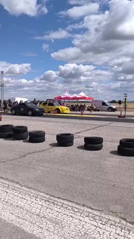 A drag race is about to begin. Two cars, one black and one yellow, are lined up side by side on a paved track. Several people stand behind a barrier of tires. Spectators are visible in the background, along with tents and a light pole with a Christmas tree light setup. The sky is blue with white clouds.