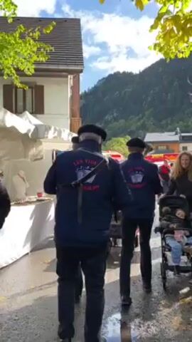 The video shows three men in matching dark jackets and berets walking through a market. They are playing musical instruments, one a wind instrument of some kind, and the other two what appears to be some kind of percussive instrument on a small wheeled cart. Behind them, people walk by, some with strollers. Market stalls are visible on the left, and in the distance, a mountain range is seen on a sunny day.