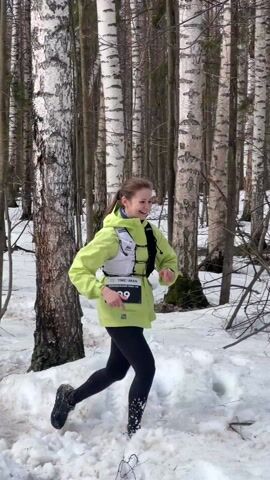 Two people are running in a snowy birch forest. The person in front is a woman with a lime green jacket and black leggings. Behind her is a man wearing a black jacket and black beanie, with a number 675 pinned to his front. He is holding a phone and appears to be looking at it. Both are running through the snow-covered forest, with many birch trees visible in the background. The man is calling to the woman to "бутель давай", give me the bottle. Another person, an older woman is visible in the background.
