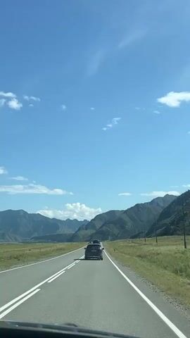 The video shows a view from the dashboard of a car driving on a paved road. The road has double white lines on the left side and a single white line on the right. The road curves slightly to the right. There are two dark-colored SUVs ahead, driving in the same direction. The SUV in front has a roof rack. To the right of the road, there is a grassy field with some trees and scattered poles. In the background, there are majestic mountains with green slopes and some rocky outcrops. The sky is a clear, vibrant blue with scattered white clouds. The sun is shining, casting shadows on the road. The overall scene depicts a scenic road trip through a mountainous region on a clear day.