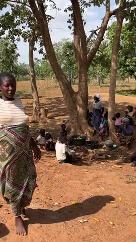 The video presents a group of people, mostly children, gathered under the shade of trees on a sunny day. The ground is reddish-brown dirt. A woman in a striped shirt and patterned skirt stands in the foreground, looking at the camera. Behind her, the group of people appears to be sorting or preparing food, as evidenced by the presence of buckets, bowls, and what seem to be fruits scattered around. Some individuals are seated, while others stand and interact. The environment is rural, with open fields and trees visible in the background.
