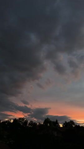 The video shows a sunset over a silhouette of trees and a house. The sky is filled with dark clouds tinged with pink and orange hues, indicating the sun is low on the horizon. The trees are dark against the colorful sky, and a small house is visible among them. The camera pans slightly to the right, revealing more of the sunset and the landscape.