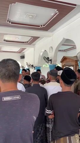 A group of men are gathered in what appears to be a covered outdoor area or a mosque. Most of the men are wearing Islamic caps (kufis) and traditional sarongs. The men are facing inwards, some are talking to each other, and some are looking towards a central point where a man is speaking or leading a prayer. There are ceiling fans visible, and the architecture suggests a warm climate. The overall atmosphere appears to be respectful and communal.