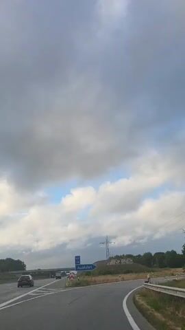 The video shows a POV shot from a vehicle driving on a road. The road appears to be a highway or a major road with two lanes. There are a few cars visible in the distance. On the right side of the road, there are trees and a power line. On the left side, there is a signpost with an arrow pointing to the right and the word "Ausfahrt" which means "Exit" in German. The sky is cloudy with some patches of blue sky. The weather appears to be overcast.