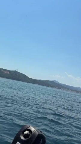 This is a first-person perspective video of a boat moving through the water. The water is blue and choppy, with small waves. In the background, there is a coastline with hills and trees. The sky is clear and blue. The video shows the back of the boat with a black engine. There are no people visible in the video. The time stamp in the video is 00:00 to 00:04.