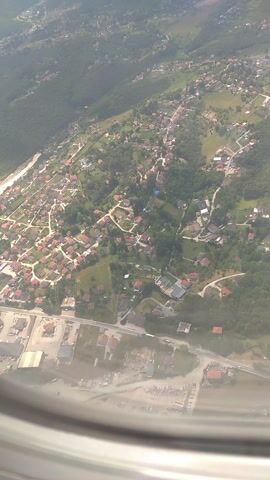 This video captures an aerial view of a town, likely Ilidža, Bosnia and Herzegovina, as seen from an airplane window. The scene is dominated by a dense collection of houses with red tile roofs, nestled among green trees and winding roads. The architecture suggests a suburban or semi-rural environment.

To the right of the residential area, there appears to be a more developed industrial or commercial zone with larger buildings, parking lots filled with vehicles, and what might be construction activity. This area seems to have a dirt or gravel surface, contrasting with the greenery and rooftops. Further to the right, the view is obscured by the airplane's wing and window frame, with some hazy atmospheric conditions visible.

The perspective is from a high altitude, making it difficult to discern specific details about the inhabitants or any immediate activities. However, the arrangement of houses and roads indicates a populated area. The lighting suggests it is daytime, with bright sunlight casting shadows, though the exact time of day is not specified. The weather appears to be clear, allowing for good visibility of the landscape below. No text is visible in the video. The overall impression is a functional town with residential and industrial areas, viewed from a detached, elevated perspective during daytime.