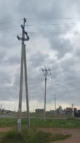 The video shows a cloudy sky and a rural landscape with power lines and poles. A car drives past on a road in the background. There is also a building and what appears to be a silo. The ground in the foreground is dirt and grass.