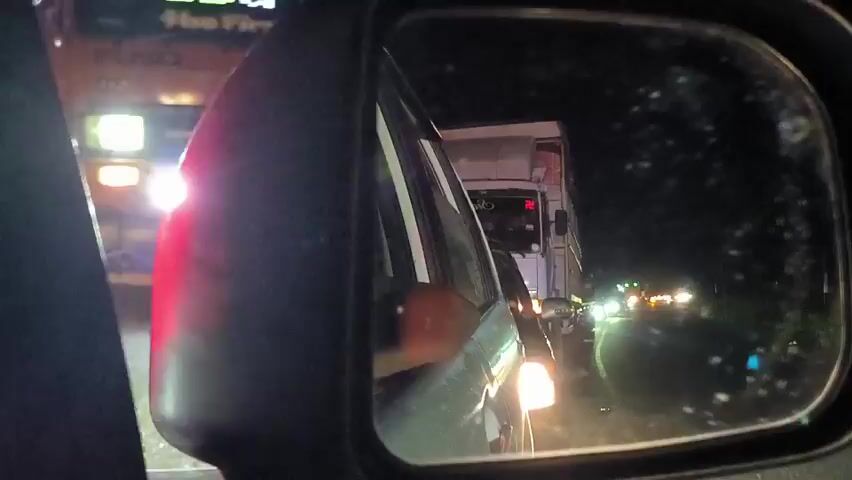 The view through a car's side mirror at night shows a line of vehicles, mostly trucks, moving slowly in traffic. The mirror is slightly water-spotted. The taillight of the car the mirror is attached to is visible in the foreground, glowing red. The other vehicles have illuminated headlights and taillights. The lights on the vehicles behind flash sporadically. The overall environment appears dark and potentially wet.