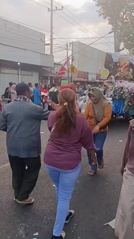 A group of people are gathered on a street, some are holding Indonesian flags. One woman in a pink and white patterned shirt is holding a flag and smiling. Another woman in a purple shirt is reaching out to her. A woman in a beige hijab is standing nearby, also smiling. The background shows buildings and a street with parked motorcycles. The scene appears to be a celebration or gathering, possibly a parade or festival.