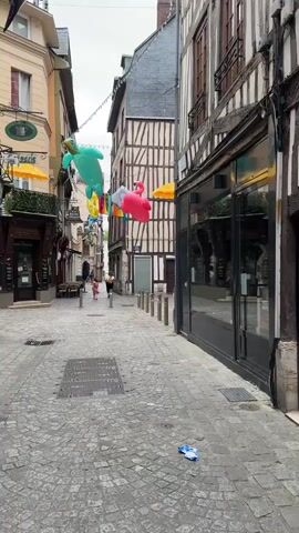 The video shows a street in a European town with cobblestone pavement and traditional half-timbered buildings. The street is decorated with colorful inflatable decorations, including sea turtles and flamingos, strung across the narrow street. In the foreground, a discarded plastic bottle lies on the pavement. The camera pans to the right, focusing on a shop window displaying two mannequins dressed in summer clothing. One mannequin wears a white sleeveless top with black polka dots and long white pants, while the other wears a strapless blue dress. The shop's name, "LE COMPTOIR des" is visible above the window, followed by "PIERRE ERZET" reflected in the glass. The display area is filled with white pebbles. To the left of this shop, another shop window is visible, reflecting the street and decorations. A person, appearing to be a child, is walking away from the camera down the street.