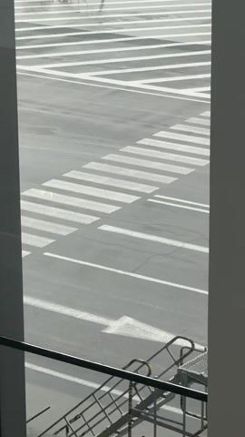 The video opens with a view of an airport tarmac from an elevated position, looking out through a large window. The tarmac is wet, indicating recent rain. White markings, including a crosswalk and directional arrows, are visible on the asphalt. In the distance, a yellow taxi with flashing orange lights on its roof drives across the tarmac, passing over the white markings. Further back, a large airplane is parked at a gate, connected to a jet bridge. Several airport personnel are visible near the airplane, some appearing to be working on the aircraft. Another airplane is visible in the background, further down the tarmac. The scene is framed by the edges of the window, with a metal railing and steps visible in the foreground, suggesting the vantage point is from a walkway or observation area.