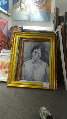 The video displays a collection of framed paintings in what appears to be an art gallery or studio. The camera is positioned low and tilted, capturing a portrait of a man in a golden frame prominently in the foreground. Surrounding this central piece are other paintings, including abstract art with koi fish, a painting of koi fish with blue and white accents, a painting of red fish, a goldfish painting, and a painting of a tiger. The paintings are stacked and leaning against each other. The floor is a dark gray tile. A person's legs and feet, wearing white sneakers with gray accents, are visible on the right side of the frame, suggesting someone is present in the space. The lighting is bright, illuminating the artwork.