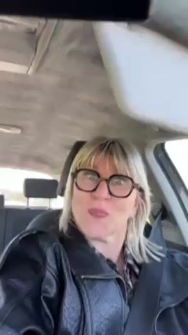 A woman with blonde hair, wearing a black leather jacket and glasses, sits in the driver's seat of a car. She is wearing a seatbelt. Initially, she makes a face at the camera, then smiles. The car appears to be in motion, as scenery passes by outside the window. The interior of the car is visible.