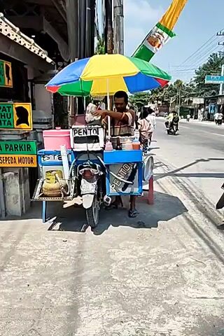 A street vendor stands by their cart, a motorcycle is parked on the right side of the road, and people are walking and riding motorcycles in the distance. The vendor's cart is decorated with a colorful umbrella. The scene is in a town or city. The sun is shining, and the sky is blue.
