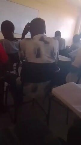 The video shows a classroom setting with students seated at desks. The camera focuses on the lap of a person sitting in a chair, wearing a dark gray skirt. Other students are visible in the background, some looking towards the front of the room. The floor is tiled.