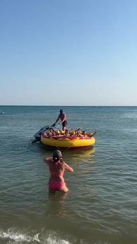 A woman in a pink bikini stands in shallow water, holding a phone up to take a picture of a yellow inflatable tube being pulled by a jet ski. On the tube are several people, including children. A man stands on the tube, holding a rope attached to the jet ski. The water is calm and blue, and the sky is clear. The sun is shining.