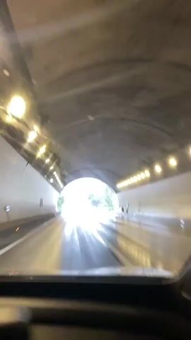 The video begins inside a tunnel as a car drives towards a bright exit. The tunnel is illuminated by overhead lights on both sides. As the car approaches the exit, the view shifts to outside the tunnel, revealing a wet road and lush green foliage. It appears to be raining, as evidenced by the raindrops on the windshield and the wet surface of the road. Traffic signs are visible in the distance, indicating an upcoming intersection or highway split. The video continues to show the car driving on the wet road under an overcast sky. The camera remains focused on the road ahead, with the surrounding environment blurred due to the movement of the vehicle and the rain.