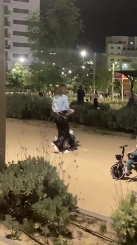 The video depicts a nighttime scene in an urban park in Kiryat Motzkin, Israel. Several people, including adults and young children, are present on a light-colored path. A young child, wearing a patterned shirt and cap, is seen kneeling and playing with white cylindrical objects, possibly bottles or cans.

An adult woman, dressed in a light blue jacket and black pants, initially stands near the playing child. A group of people then walks past, including a man in a white sweater pushing a stroller containing a child, and a woman in a light blue top walking alongside him. Another child is visible riding a small tricycle. Following this, the woman in the light blue jacket kneels down to engage with the child who is playing on the ground. The background features illuminated park areas, planted bushes, and multi-story residential buildings, indicating an urban setting after dark. No discernible text is visible.