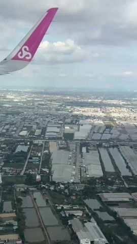 The video is an aerial view from an airplane looking down at an industrial area. The airplane's pink wingtip with a white logo is visible in the upper left corner. The landscape below consists of numerous large, rectangular buildings, likely warehouses or factories, organized in rows. Interspersed between these buildings are bodies of water, possibly canals or ponds, and patches of greenery. In the distance, a hazy cityscape with more buildings and a faint mountain range can be seen under a partly cloudy sky. An automated voice announces that the airplane will be landing soon and instructs passengers to ensure their seatbelts are securely fastened.