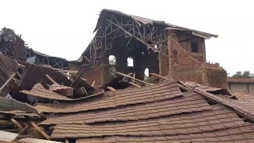 The video shows a building heavily damaged. The building is made of brick and has a roof made of brown tiles. The roof is partially collapsed, and there are piles of debris on the ground, including broken bricks, wood, and metal. The building appears to be old and in disrepair. The sky is overcast.