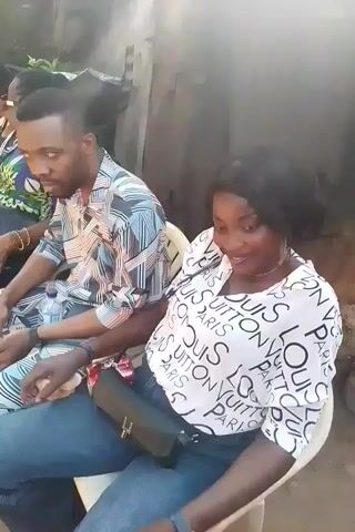 The video shows a group of people sitting on chairs outdoors. The people are of various ages and genders, with the majority appearing to be women wearing colorful dresses with floral patterns. A few men are also present, wearing patterned shirts and casual clothing. They are sitting in a line and looking in different directions, some are looking at their phones and some at each other. In the background, there is greenery and a glimpse of a building. There are drinks on a table and a bucket in the background.