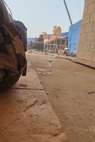 The video shows a partially obscured view of a movie set, likely outdoors and under bright sunlight. In the foreground, there's a large bag, possibly a backpack, obscuring much of the left side of the frame. The ground is a dusty, sandy color, littered with debris including two empty water bottles. Further in the background, a man is visible sitting on a structure made of wooden scaffolding. On top of the scaffolding are two golden statues that resemble griffins. Behind the scaffolding, a blue backdrop is partially visible. Other people are moving around in the distance, but they are out of focus and difficult to discern. The overall impression is that the video was taken surreptitiously from a hidden position, focusing on the immediate foreground while capturing the movie set in the background.