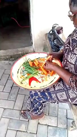 An eye-level outdoor shot shows a person sitting and peeling carrots onto a round tray. The person, who appears to be a woman, is seated on what looks like a wooden bench and has the tray in her lap. The tray contains peeled carrots, green onions, and a red pepper. The woman is wearing glasses, a patterned top, and patterned pants. She is also wearing sandals. Next to her is a black plastic bag and a metal container on the bench. The ground is paved with grey bricks. Behind her is a doorway leading into a dark room with various items on a shelf. The camera moves slightly during the shot. A logo appears at the bottom left of the frame.