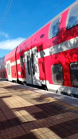A red and white train pulls away from a train station platform on a sunny day, passing the camera as it recedes into the distance. The camera pans up to show the engine as the train speeds away. The clock at the station reads 11:56.