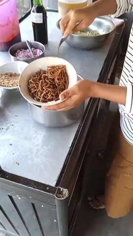 A person's hands are visible preparing food at a metal counter. The person is holding a bowl of noodles and placing it on the counter next to other bowls containing ingredients like chopped red onions, crispy fried shallots, and chopped green onions. A large metal bowl of noodles sits in the center, and a fork is placed in it. In the background, there are containers of red sauce and what appears to be a bottle of dark liquid. The environment suggests an outdoor food stall or a simple kitchen setup. The person's hands are the primary focus, interacting with the food and utensils.