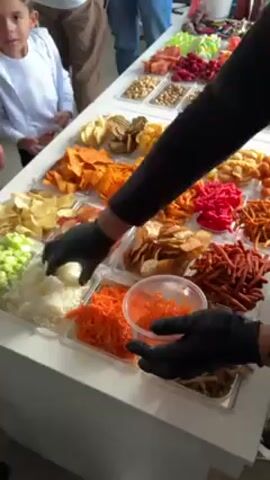 The video shows a food preparation scene. A person wearing black gloves is combining chopped onions and another ingredient in a plastic container. This container is one of many set out on a white table, containing various food items. The table displays an assortment of colorful snacks, including chips, shredded carrots, chopped celery, and other unidentified ingredients. Several people, including a young boy in a white shirt, are standing around the table, possibly waiting for the food to be prepared. The overall setting seems to be an event or gathering where snacks are being prepared for guests.