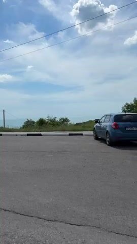 The video opens with a low-angle shot of an asphalt road with cracks, revealing a dirt verge to the immediate right. Two white cars are parked side-by-side on the right side of the road, a Kia Soul on the left and a larger SUV on the right, both facing away from the camera. Beyond the cars, a grassy hill slopes down towards a visible body of water under a partly cloudy blue sky. As the camera pans to the right, the cars move out of frame, revealing a white, sail-shaped structure on the grassy hillside. The pan continues, showing more of the green, shrub-covered landscape, a utility pole, and the distant water under the vast sky. The foreground remains a mix of dirt and sparse vegetation. The sound of wind or vehicle movement is audible throughout the clip.