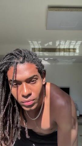 Here is a detailed description of the video:
The video features a young, shirtless man with dark skin and dreadlocks. He is wearing a silver chain. He is standing in a brightly lit room with white walls and a white ceiling.  Sunlight streams in through a window, casting shadows across the ceiling. In the background, there is a television, a wall clock, a white table, and a white electric kettle. The man looks directly at the camera, and touches his hair. There is music playing in the background.