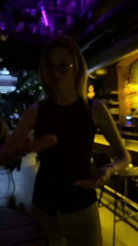A woman with fair skin and blonde hair stands in the center of the frame, in what appears to be a dimly lit bar or club. She is wearing glasses, a black sleeveless top, and light-colored pants. She is facing the camera and making gestures with her hands, moving them in a circular, spinning motion in front of her chest.  The environment is filled with the soft, hazy light of the club. Behind the woman is a bar area with stools, a brick wall, and shelves with bottles. The overall lighting is a mix of yellow and purple hues. Other people can be seen in the background, although they are out of focus.