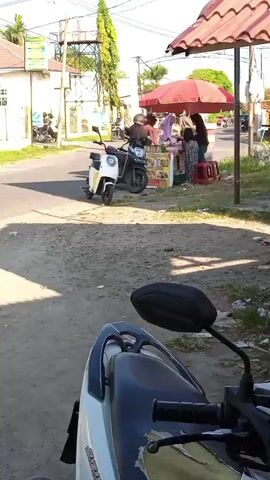 A still shot captures a street scene. In the foreground, the back of a white and blue motorcycle is visible. To the left, a white electric scooter and a dark motorcycle are parked on the side of a road. Further down the road, a food stall with a red umbrella is set up. Three individuals are standing at the stall, two appearing to be customers and one serving. A person on a motorcycle is visible in the background on the left. Buildings and a water tower are also present in the environment. The scene appears to be outdoors, under daylight.