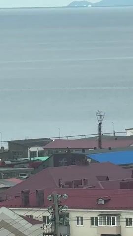 A distant view shows a wide expanse of gray water under an overcast sky. In the mid-ground, a small white boat speeds across the water, leaving a foamy wake. Below the water, a coastal town with red-roofed buildings is visible. A tall metal tower stands on the right side of the frame, and a few other industrial-looking buildings are scattered among the residential structures. In the far distance, hazy outlines of islands or mountains can be seen on the horizon. The overall impression is of a somewhat industrial or working waterfront under a subdued, cloudy sky.