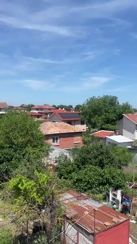 The video shows an aerial view of a rural village on a sunny day. The sky is blue with some scattered white clouds. The foreground shows a dirt road and a small red garage with a rusty metal roof. Behind the garage, there are several houses with terracotta roofs, surrounded by lush green trees and vegetation. Some houses have solar panels installed on their roofs. In the background, more houses and trees are visible, extending to the horizon. The overall impression is that of a peaceful, residential area in a warm climate.