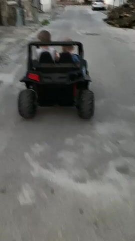 Two children are riding in a black ride-on toy ATV on an asphalt street. The child in the driver's seat appears to be older than the child in the passenger seat. The ATV has large black tires. The street is lined with buildings and a pile of chopped wood is visible on the right side of the street. The sky is overcast.