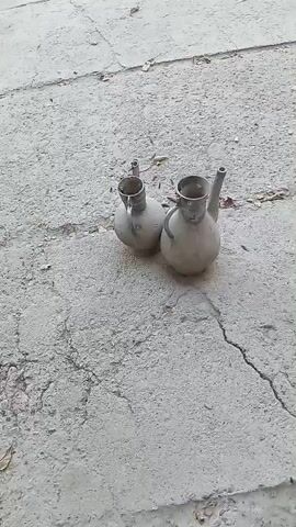 The video shows two light-colored, bulbous pitchers with narrow spouts and handles resting on a cracked concrete surface. The pitchers appear to be made of metal and have a somewhat aged or worn appearance. The concrete is a pale gray and shows significant cracks, with some dried leaves or debris scattered on it. The lighting suggests it is daytime, possibly outdoors.