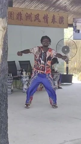 A man with dreadlocks is dancing in front of a wooden sign with Chinese characters. He is wearing a colorful patterned shirt and blue pants with a similar pattern. His arms are outstretched and his body is angled to his right. There is a large fan behind him, and to his left, several drums are visible. Another person can be seen in the background, also appearing to be involved in the performance. The overall setting appears to be an outdoor stage or performance area, possibly at a cultural attraction.