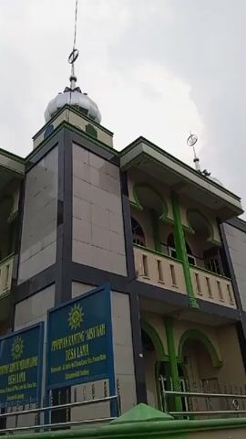 A low-angle exterior shot shows a multi-story building with a green and beige facade, featuring Islamic architectural elements such as arched windows and decorative trim. The building has a prominent dome topped with a crescent moon symbol. Two blue signs with yellow emblems are mounted on the front, one partially obscured, with text in what appears to be Indonesian. A green and white fence runs along the front of the property. The sky is overcast. The video pans slightly to the right.