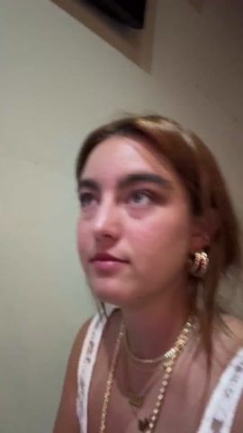 A young woman is featured in a close-up, selfie-style video, positioned against a plain, light-colored wall. She has medium-length, light brown hair with visible golden highlights and prominent dark eyebrows. She is wearing multiple layered gold chain necklaces, one of which has a small pendant, and large, thick gold hoop earrings. Her top appears to be a white lace or crocheted camisole.

The woman is speaking, her mouth moving as she articulates the words "Absolutely exhausted." Despite her statement, her facial expression is conversational and relatively neutral, occasionally glancing slightly upwards and to the side. In the upper background, a dark, rectangular object, possibly a flat-screen television or monitor, is partially visible. Upbeat electronic or dance music is distinctly audible in the background, suggesting a lively indoor environment. The lighting is artificial and appears somewhat warm. No visible text is present in the frame.