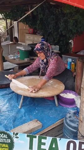 A woman wearing a headscarf and a patterned pink shirt is rolling out dough on a round wooden surface supported by two small legs. The surface is covered in a blue tarp. There is a bowl of white powder, likely flour, to the right of the woman. Behind her, there are shelves with various items, including bottles and containers, and some greenery hangs from above. To the left, there is a glimpse of another person and some items on a table. In the foreground, a sign with green text and a leaf logo is partially visible, with the words "DOĞAL LEZZETLER" and "NATURAL FLAVORS" appearing on it. The overall setting appears to be an outdoor food stall or market.
