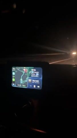 The video shows a car driving on a dark highway at night. The dashboard is visible, displaying a navigation system with a route mapped out and music playing. The road ahead is illuminated by the car's headlights, and a few distant lights are visible. The overall scene is dark and focused on the act of driving through the night.