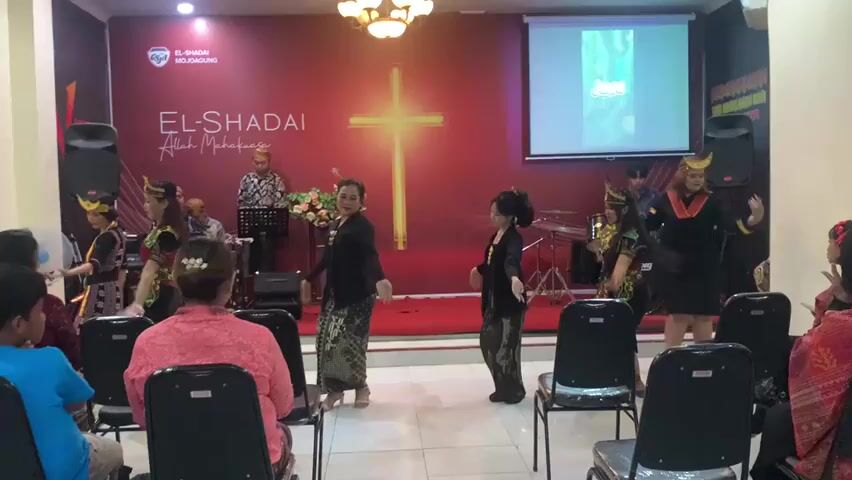 This is a video recording of a performance taking place indoors, likely in a church or community hall. The background features a large red banner with a logo that reads "EL-SHADAI ALLAH MAHA KASIH" and a prominent illuminated cross. To the right of the cross, a projector screen displays an image with the word "Yesus" in text.

In the foreground, facing the audience, are several individuals performing a choreographed dance. Three women in traditional attire are at the center, with two young girls in similar outfits to their left. Another performer in a black dress with yellow accents is to the right, and a woman in a black outfit with a red and yellow patterned sash is further right. They are all engaged in various dance movements.

Behind the performers, on a raised platform, a man in a patterned shirt is standing near a keyboard and a microphone stand. Further back, a drum set is visible.

The audience is seated in chairs, facing the stage. Some audience members are visible in the foreground and to the sides, including a person in a blue shirt on the left and individuals in pink and patterned clothing on the right. The overall environment appears to be a well-lit, indoor space with white tiled flooring.
